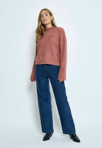 Rosa, flauschiger Strickpullover mit hohem Kragen und langen, leicht ausgestellten Ärmeln, kombiniert mit weit geschnittenen blauen Jeans und schwarzen spitzen Stiefeln.