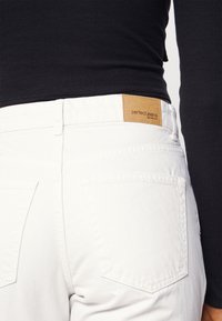 Jeans brancos com um tecido texturizado, apresentando bolsos traseiros e uma etiqueta de couro castanho marcada com "perfect jeans" perto da cintura.