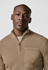 Beige gerippter Strickpullover mit einem halben Reißverschluss und einer Brusttasche; strukturierter Stoff mit lässiger Passform.