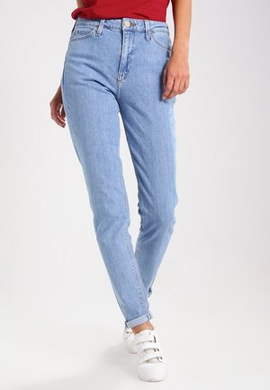 Persoon draagt lichtblauwe high-waisted jeans met opgerolde boorden, gecombineerd met witte klittenbandsneakers en een rood topje, staand op een witte achtergrond.