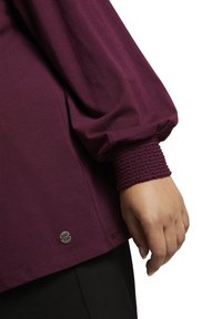 Main portant un haut à manches longues violet avec un poignet texturé et un petit logo métallique rond près de l'ourlet, sur un fond blanc.