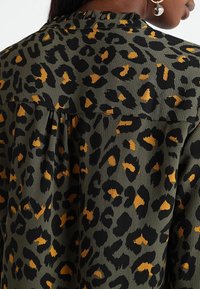 Grüner Stoff mit einem schwarzen und orangenen Leopardenmuster, weicher Textur und mit Raffung am Ausschnitt und an den Nähten.