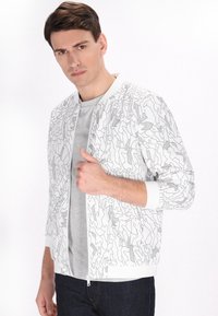 Witte bomberjack met zwart abstract lijnpatroon, geribde manchetten en zoom, ritssluiting aan de voorkant, gedragen over een grijs t-shirt en donkere spijkerbroeken.