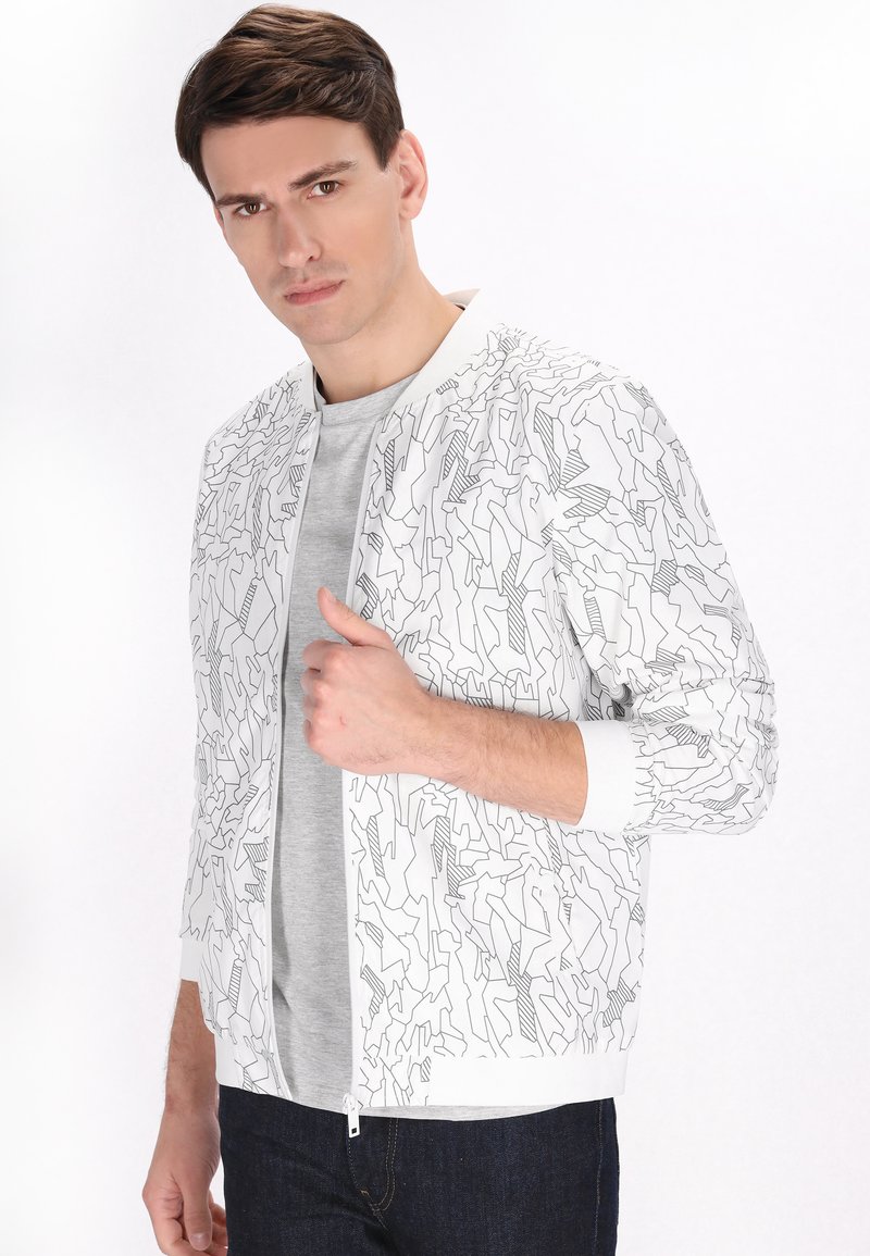 Witte bomberjack met zwart abstract lijnpatroon, geribde manchetten en zoom, ritssluiting aan de voorkant, gedragen over een grijs t-shirt en donkere spijkerbroeken.