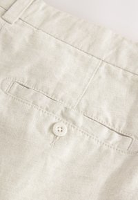 Pantalon en coton beige clair avec une poche arrière et un bouton blanc. Le tissu a une texture lisse avec des lignes de couture visibles.