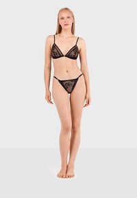 Conjunto de lencería de encaje negro que incluye un bralette triangular con tirantes finos y un tanga a juego. La tela es translúcida con patrones florales.
