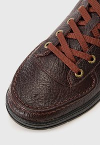 Sneaker in pelle marrone scuro con superficie testurizzata, occhietti in bronzo e lacci marroni. Presenta una punta arrotondata e dettagli di cucitura a contrasto.