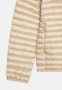 Beige gewatteerde jack met een glanzende textuur, voorzien van horizontale stiksels en een ritszak aan de zijkant. Soepele, lichte stof.