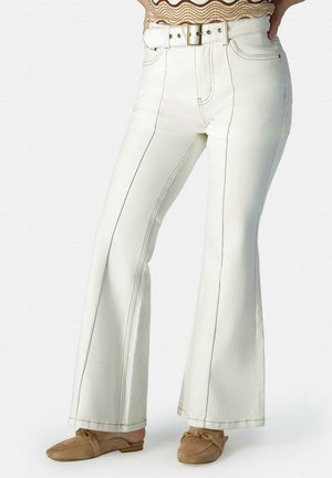 Witte, hooggetailleerde flared broek met zichtbare stiknaden, gecombineerd met bruine instap loafers en een patroonrijke top die is ingestopt.