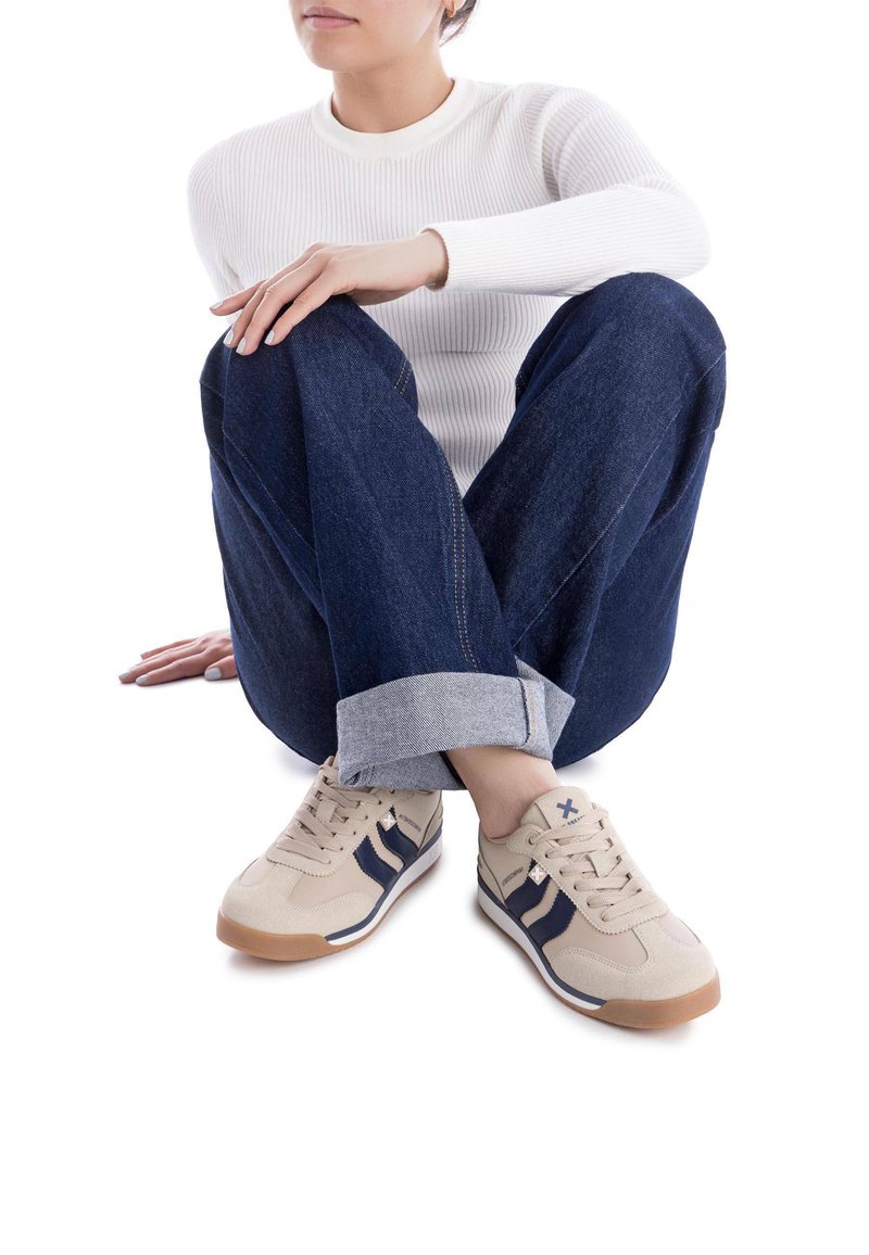 Zapatillas deportivas beige con detalles en azul marino, material de ante, suela de goma y diseño de cordones, llevadas con jeans de mezclilla oscura remangados.