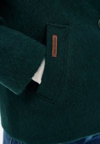 Kieszeniowy, ciemnozielony, teksturowany wełniany płaszcz z brązową metką "blue shadow" oraz białym materiałem częściowo widocznym wewnątrz kieszeni.