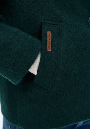 Donkergroene, textuurrijke wollen jaszak met een bruine 'blue shadow'-label en een gedeeltelijk zichtbaar wit stuk stof binnenin de zak.