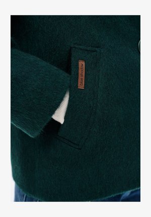 Donkergroene, textuurrijke wollen jaszak met een bruine 'blue shadow'-label en een gedeeltelijk zichtbaar wit stuk stof binnenin de zak.