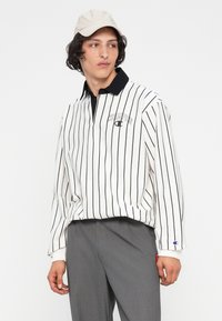 Bílo-černý pruhovaný polo s límcem na zip a modrou logem, v kombinaci se šedými kalhotami. Tkanina působí hladce a má ležérní styl.