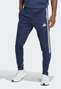 Die Marineblauen Adidas Trainingshosen haben eine schmal zulaufende Passform, einen elastischenBund und weiße Three-Stripes-Akzente an den Seiten. Sie bestehen aus glattem Material.
