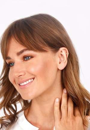 Goldener Creolenohrring, der am oberen Ohr getragen wird und ein minimalistisches Design aufweist. Die Haare sind braun und gewellt, was den eleganten Stil des Ohrrings ergänzt.