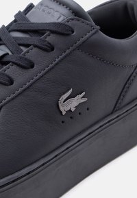 Μαύρο δερμάτινο αθλητικό παπούτσι με μαύρα κορδόνια, χοντρή σόλα και ασημένιο λογότυπο κροκόδειλου στην πλευρική επιφάνεια.