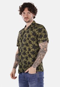 Camicia a maniche corte con bottoni in tessuto verde oliva, presenta un motivo di palme nere, vestibilità rilassata e colletto aperto.