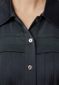 Chemise noire avec un col pointu, présentant une patte de boutonnage avec trois boutons, un tissu texturé et de subtils plis verticaux.
