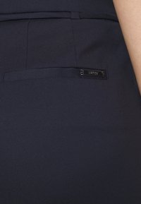 Detailní záběr na tmavě modré kalhoty s malou zadní kapsou a kovovou značkovou visačkou s nápisem "Esprit" na pase.