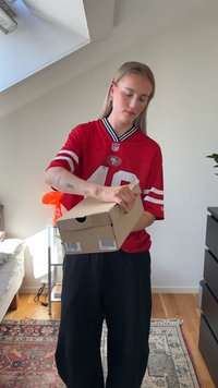 Persoon in een rood voetbalshirt opent een kartonnen doos in een lichte kamer met een gemarmerd tapijt en meubels.