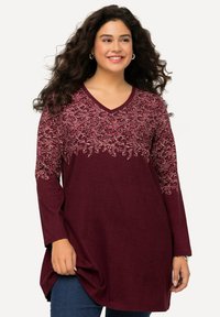 Frau mit lockigem Haar, die lächelt, trägt eine langärmlige bordeauxfarbige Tunika mit rosa floralem Stickereimuster und blaue Jeans, steht vor einem schlichten Hintergrund.