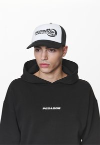 Fekete, laza fazonú kapucnis pulóver, a mellkasán nagy, fehér "PEGADOR" logóval, amelyhez egy fekete-fehér trucker sapka társul, rajta a "PEGADOR ADVENTURES" felirattal.