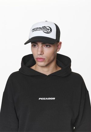 Crna dukserica s širokim krojem i velikim bijelim logotipom "PEGADOR" na prsima, u kombinaciji s crno-bijelim trucker kapom koja nosi natpis "PEGADOR ADVENTURES."