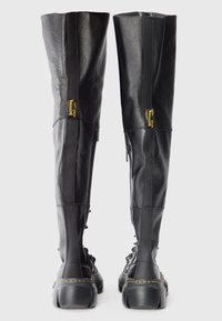 Une paire de bottes noires en cuir montant, à lacets, avec des fermetures éclair sur les côtés et des semelles épaisses, vues de l'arrière sur un fond blanc.