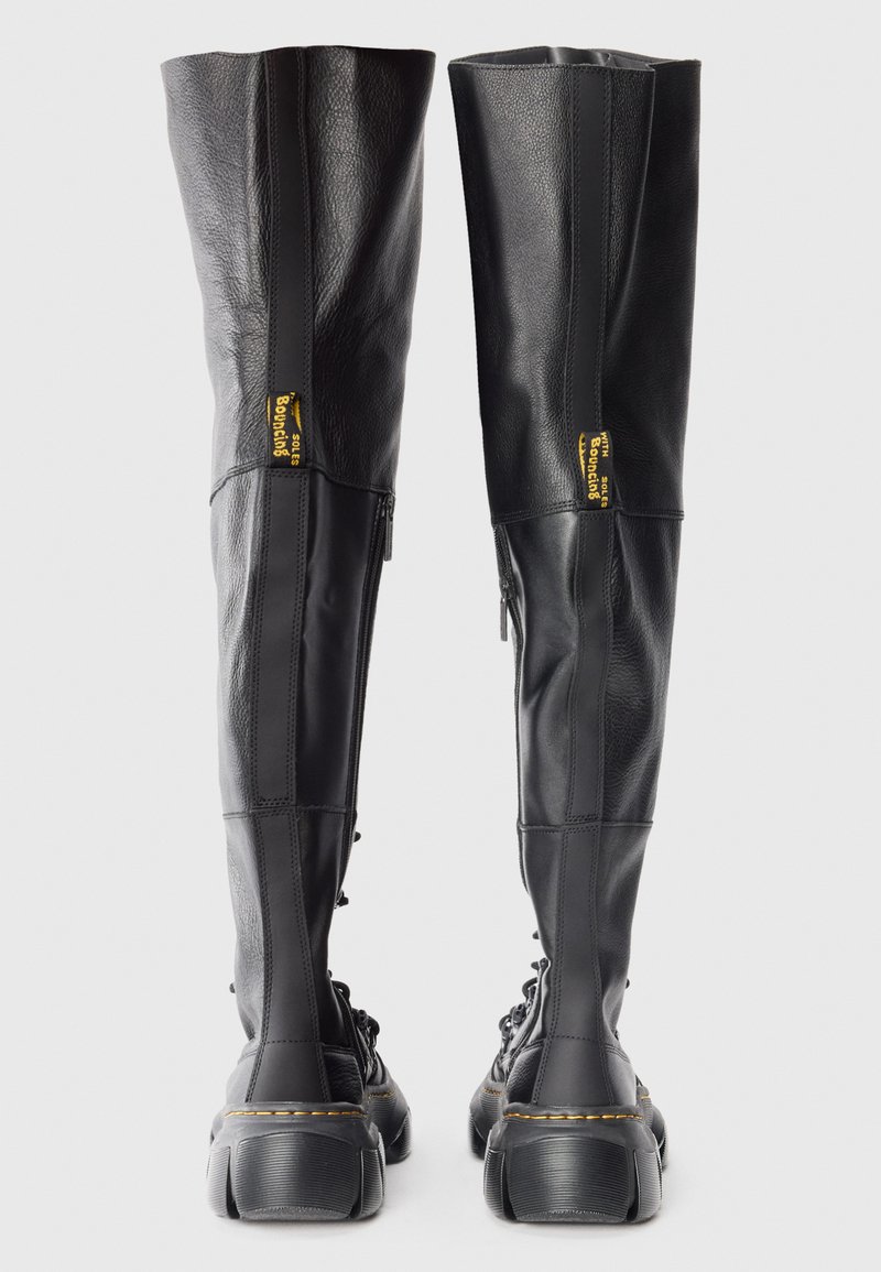 Un paio di stivali alti neri in pelle con lacci, zip laterali e suole spesse visti da dietro su uno sfondo bianco.