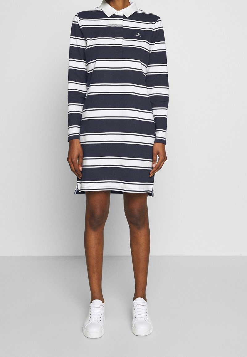 Gestreiftes Polo-Kleid in Marineblau und Weiß mit langen Ärmeln, einem Kragen und seitlichen Schlitzen, kombiniert mit weißen Sneakers. Material aus Baumwollmischung.