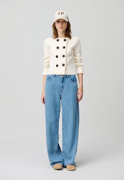 Maglione a coste crema con bottoni neri, abbinato a jeans a gamba larga azzurri e scarpe slip-on beige, completato da un cappellino beige.
