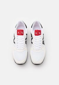 Scarpe sportive bianche con accenti neri, rete testurizzata e pannelli lisci, lacci piatti e una linguetta rossa con il marchio "A|X."