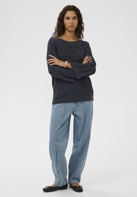 Marineblauer, gestrickter Pullover mit Wellenmuster, weiten Ärmeln, kombiniert mit hellblauen, locker sitzenden Jeans und schwarzen Ballerinas.