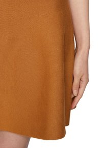 Robe en tricot couleur rouille avec une texture lisse, un design évasé et une longueur au-dessus du genou, avec une main posée sur le vêtement.
