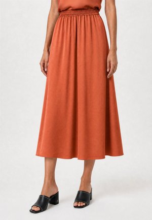 Mujer con falda midi de talle alto color óxido con cinturilla elástica, combinada con sandalias negras de tacón bloque y punta abierta.