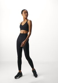 Črn športni crops top in visokorasle legice, oba v rebrasti teksturi. Model nosi črne tekaške čevlje, stoji na beli podlagi.