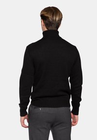 Maglione nero a collo alto in maglia, con polsini e orlo a coste, caratterizzato da una texture liscia e un design aderente, indossato con pantaloni grigi su misura.