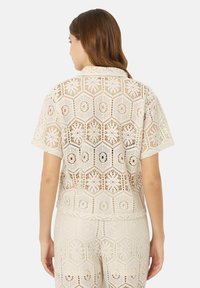 Blusa de encaje crema de manga corta con intrincados recortes hexagonales, que muestra una tela ligera y texturizada. Vista trasera con cuello liso.
