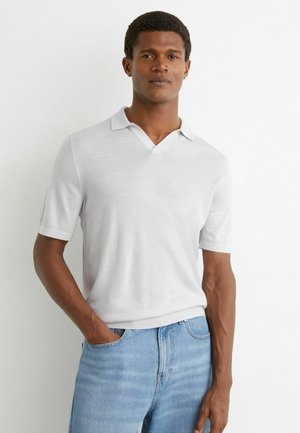 Mand med kort krøllet hår iført en hvid kortærmet polo og lyseblå jeans, stående med den ene hånd i lommen.