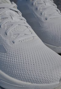 Zapatillas deportivas blancas con parte superior texturizada que presenta pequeñas perforaciones, cordones planos y una suela acolchada. Diseño sencillo y mínima marca.