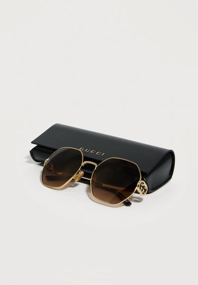 Καφέ γυαλιά ηλίου με εξαπλάσια καφέ φακούς και χρυσό πλαίσιο, ακουμπισμένα σε μια μαύρη θήκη με το λογότυπο Gucci πάνω σε λευκή επιφάνεια.