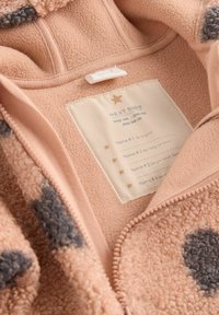 Fleece-babyjacka i mjuk persikofärg med gråa prickar. Har dragkedja framtill och en vårdtagg med plats för namn.