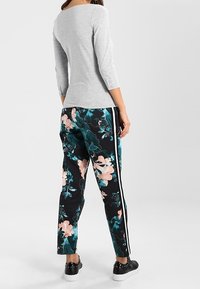 Grauer Oberteil mit drei Viertelärmel, floral gemusterte schwarze Hose mit türkisfarbenen und rosafarbenen Blumen, weißem Seitenstreifen und schwarzen Sneakers.