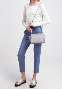 Mujer vestida con un suéter blanco abotonado, pantalones cortos azules, sandalias deslizantes metálicas y sosteniendo un bolso cruzado gris con tachuelas.