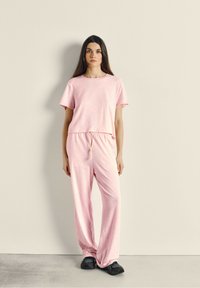 Ensemble de vêtements de détente rose comprenant un t-shirt ample et un pantalon élastique à cordon de serrage. Tissu doux au confort relaxé, associé à des baskets noires.