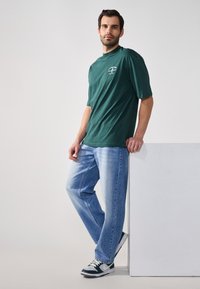 Grünes Baumwoll-T-Shirt mit hohem Kragen und kurzen Ärmeln, kombiniert mit hellblauen Jeans und dunklen Sneakern, stehend vor einem weißen Block.
