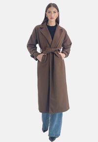 Bruine trenchcoat met ceintuur, reverskraag, lange mouwen en zijzakken, gecombineerd met lichtblauwe wijde jeans en zwarte puntige hakken.