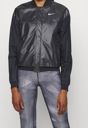 Person trägt eine schwarze Nike-Jacke mit Reißverschluss und graue gemusterte Leggings, steht entspannt mit den Armen seitlich an einem schlichten Hintergrund.