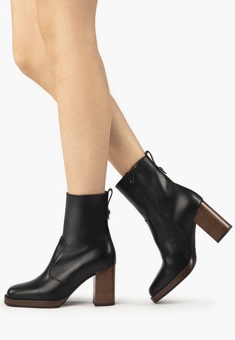 Jambes portant des bottines en cuir noir à talons blocs en bois avec fermetures éclair latérales, un pied légèrement levé sur un fond blanc.